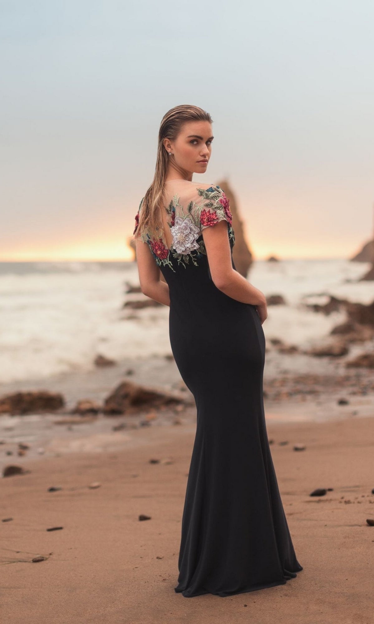 Wearing the Mac Duggal 11817 Floral Long Black Formal Dress, a woman stands on a beach at sunset, gazing over her shoulder toward the camera.